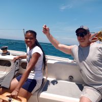 Combo Course- Yacht crew & day Skipper:This image is showing a confident student at the helm of a sailing yacht, navigating through clear blue waters under the watchful eye of an experienced instructor. The student demonstrates their newfound knowledge of navigation and seamanship. The sparkling ocean and clear blue sky provide a picturesque backdrop as they share laughter.